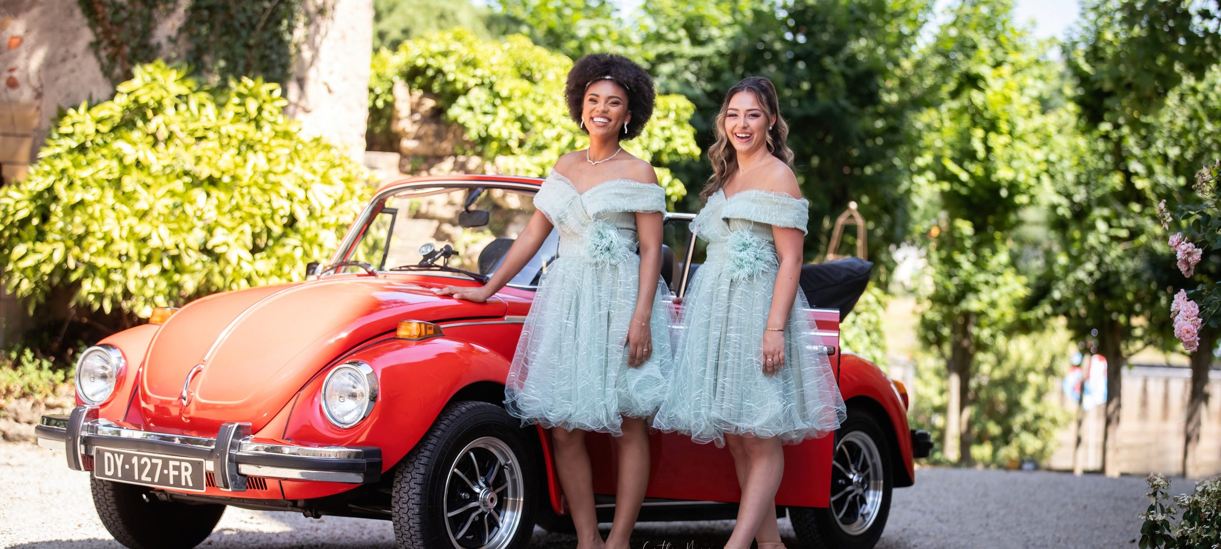 Deux demoiselles d'honneur en robes bleu pâle devant une voiture ancienne rouge